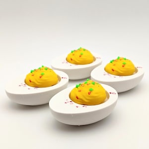 May include: Four ceramic deviled eggs, each with a bright yellow filling and green and red sprinkles. The eggs are white and sit on a white surface. A food-themed decorative item.