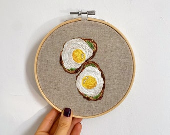 May include: Embroidered artwork of two slices of toast with fried eggs on top, displayed in a wooden embroidery hoop.
