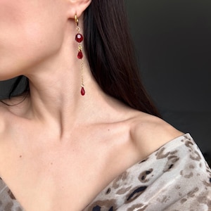 Peut inclure: Boucles d'oreilles créoles dorées avec une chaîne pendante et des perles de cristal rouges.