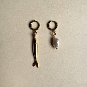 May include: A pair of gold-colored earrings. One earring features a fish pendant, while the other has a white, irregular-shaped pearl. Both earrings have a small hoop design for the ear.