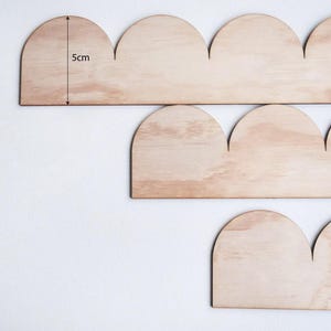 May include: Wooden cloud-shaped decorative pieces. The pieces are made of light-colored wood and are approximately 2 inches tall. The image shows several pieces arranged in a staggered pattern against a white background. The pieces are likely for home decor.