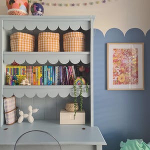 May include: A light blue desk with a scalloped bookshelf, featuring three orange gingham storage bins. The shelf holds colorful books and decorative items. A framed floral print hangs on a blue wall, with a clear chair in front of the desk.