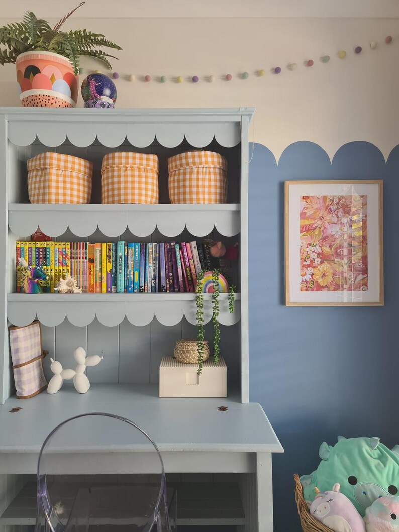 May include: A light blue desk with a scalloped bookshelf, featuring three orange gingham storage bins. The shelf holds colorful books and decorative items. A framed floral print hangs on a blue wall, with a clear chair in front of the desk.