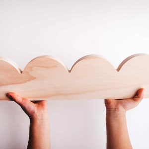 May include: A wooden shelf with a scalloped edge. The shelf is made of light wood and is held up by two hands.