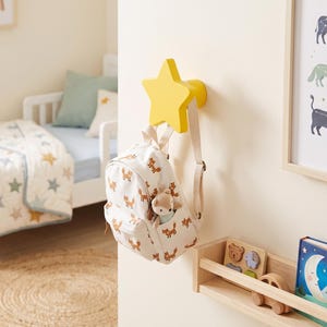 Könnte beinhalten: Ein leuchtend gelber, sternförmiger Wandhaken an einer cremefarbenen Wand. Ein kleiner, cremeweißer Rucksack mit einem sich wiederholenden orangefarbenen Fuchsmuster hängt an dem Haken, mit einem kleinen Plüschfuchs in der vorderen Reißverschlusstasche.