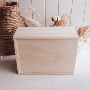 May include: A light-coloured wooden box with a hinged lid. The box is rectangular and has a natural wood grain finish. The background includes a woven basket and dried floral arrangements.