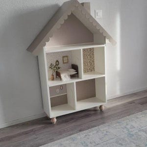 May include: A white dollhouse with a house-shaped roof and scalloped trim. The interior features miniature rooms with wallpaper, furniture, and decorative items. The dollhouse is elevated on small, rounded feet.