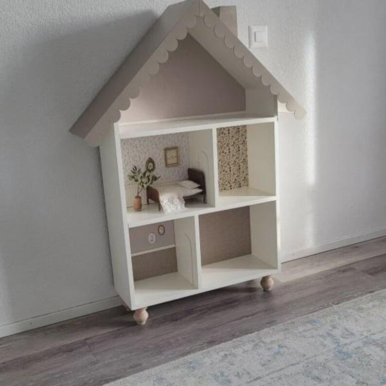 May include: A white dollhouse with a house-shaped roof and scalloped trim. The interior features miniature rooms with wallpaper, furniture, and decorative items. The dollhouse is elevated on small, rounded feet.