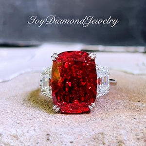 Peut inclure: Une bague ornée d'une pierre précieuse rouge vif, avec une pierre centrale de grande taille, de coupe rectangulaire. La bague est flanquée de deux pierres claires, rectangulaires, plus petites. La bague est sertie dans un anneau argenté. Le texte "Ivy Diamond Jewelry" est visible.