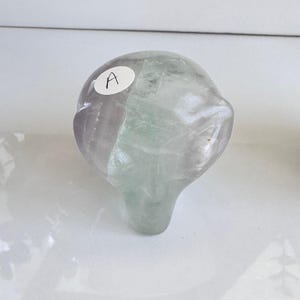 May include: A translucent, rounded stone sculpture with a light purple and green color gradient. The stone has a smooth, polished surface and a small, rounded base. A white sticker with the letter "A" is affixed to the top of the stone.