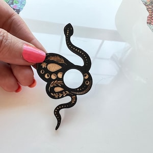 May include: A black wooden snake-shaped ornament featuring a circular cutout in the center and intricate laser-engraved details including leaf patterns, small dots, and moon phases in a natural light wood tone against the dark surface.
