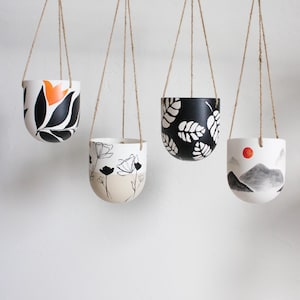 May include: Four hanging planters with different designs. The planters are white with black and orange painted designs. The planters are hanging from twine.