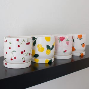 May include: Four white ceramic plant pots with matching saucers. Each pot features a different fruit design: cherries, lemons, strawberries, and oranges. The pots are arranged on a black shelf against a white wall.