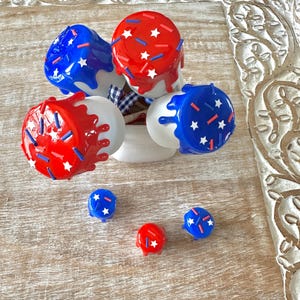 May include: A collection of red and blue decorative knobs with white bases, adorned with star and sprinkle details. The knobs are arranged on a wooden surface, evoking a patriotic theme. Some knobs have a dripping paint effect.