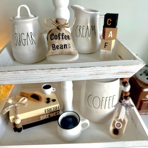 May include: A two-tiered white wooden tray with coffee-themed decor. Items include a sugar jar, a cream pitcher, a burlap bag of coffee beans, and a coffee grinder. The tray also holds a small coffee cup and a decorative block spelling "CAF".