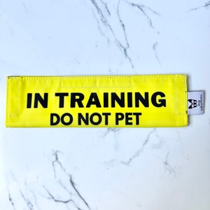 May include: Yellow dog training band with black text that reads "IN TRAINING DO NOT PET". The band has a small white tag with a black dog silhouette and the text "dog in training" on it.