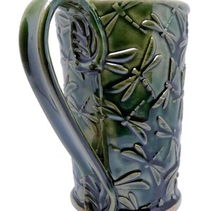 Może przedstawiać: Zielony ceramiczny kubek z wzorem ważki. Kubek ma duży uchwyt i błyszczące wykończenie. Wzór ważki jest wytłoczony na powierzchni kubka. Kubek ma około 13 cm wysokości.