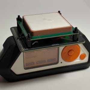 May include: A handheld electronic device with a black and white casing, featuring an orange circular navigation pad and a digital screen displaying text. A green circuit board with a square white antenna module is mounted on top with black spacers.