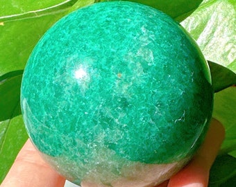 Puede incluir: Una esfera de aventurina verde, un tipo de cuarzo, con una superficie lisa y pulida. La esfera es aproximadamente del tamaño de una pelota de golf y tiene un color verde moteado.