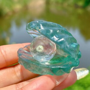 Op de afbeelding: Een heldere glazen parel ligt in een groene glazen schelp. De schelp is open en de parel is zichtbaar. De schelp is een decoratief object.