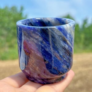 Puede incluir: Una taza de sodalita azul y blanca pulida, sostenida a mano. La taza tiene una base redondeada y una forma cilíndrica lisa. Los patrones naturales de la piedra crean una apariencia veteada única. La taza se sostiene sobre un fondo de follaje verde y cielo azul.
