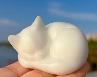 May include: A small, white, sleeping cat figurine. The cat is curled up, with its tail wrapped around its body. The figurine is smooth and glossy, with a simple, elegant design. The cat is resting on a person's hand.
