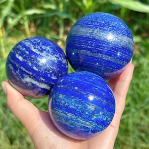 May include: Three polished lapis lazuli spheres. The spheres are a deep blue colour with streaks of white and gold. The spheres are held in a hand, with a blurred green background.