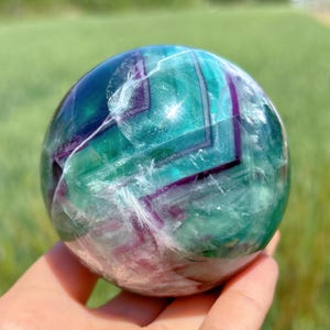 May include: A green, purple and white fluorite sphere with a polished finish. The sphere has a swirling pattern of colours and is a beautiful example of this crystal.