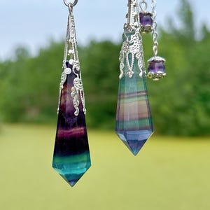 Pode incluir: Dois pingentes de pedras preciosas com tampas e correntes de filigrana prateadas. Os pingentes são alongados, facetados e exibem faixas de roxo, verde e azul. Um pingente tem uma pequena conta roxa em sua corrente.