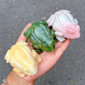 Puede incluir: Tres figuritas de rana talladas en piedra de diferentes colores. Las ranas son verdes, rosas y amarillas. Las ranas están sentadas en una mano.
