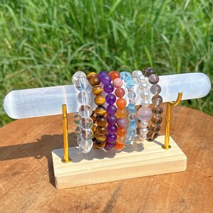 Peut inclure: Présentation de bracelets de perles de différentes couleurs, dont transparent, marron, violet, orange et bleu. Les bracelets sont disposés sur une barre en cristal de sélénite blanche, soutenue par un support en bois avec des accents métalliques dorés. Fond vert flou.
