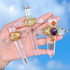 May include: Three clear quartz crystal crosses with gold accents and colorful gemstone accents. The crosses are adorned with green leaves and feature a variety of gemstones, including amethyst, carnelian, and citrine.