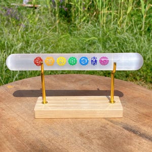 Peut inclure: Une baguette de cristal de sélénite transparente avec sept symboles de chakra colorés, présentée sur une base en bois avec des supports métalliques dorés. La baguette mesure environ 20 cm de long.