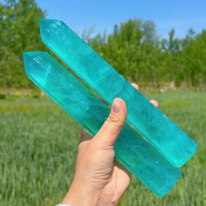 May include: Two teal blue crystal wands with a smooth, polished finish. The wands are held in a hand against a green grassy background.