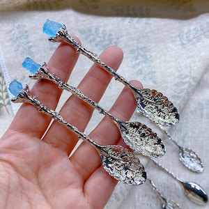 May include: Three silver spoons with ornate handles and blue crystal tops. The spoons have a leaf-shaped design on the bowl.