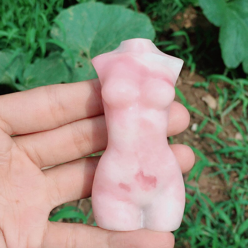 Op de afbeelding: Een roze en wit gesneden torso-beeldje gemaakt van gepolijst steen. Het beeldje heeft een glad, gepolijst oppervlak en een gedetailleerd ontwerp.
