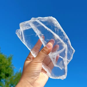 以下が含まれることがあります： 層状で半透明な外観を持つ、大きな透明なセレナイト結晶のスラブ。結晶は明るい青い空を背景に手に持たれています。