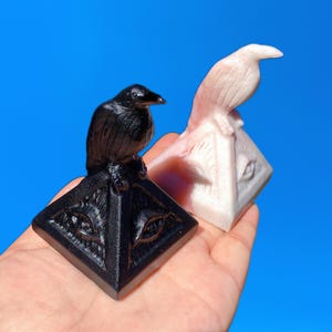 May include: Two black and white pyramid-shaped sculptures with a bird perched on top of each. The pyramids have an all-seeing eye design on the front. The bird on the black pyramid is black, and the bird on the white pyramid is white.