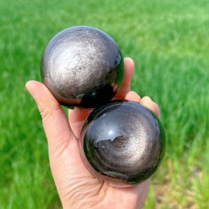Op de afbeelding: Twee zwarte obsidiaankogels met een zilveren glans. De kogels zijn glad en gepolijst en liggen op een hand.