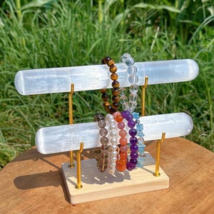 May include: A two-tiered bracelet display stand with a light wood base and gold-colored metal supports. The display holds several beaded bracelets in various colors, including brown, clear, purple, and blue. The background features green foliage.
