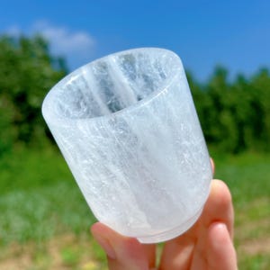 Puede incluir: Una taza cilíndrica de cristal transparente con una base ligeramente redondeada. La taza se sostiene en una mano, con un fondo borroso verde y azul. El cristal tiene un aspecto translúcido y nublado.