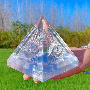 May include: A clear crystal pyramid with two eyes carved into the front. The pyramid is sitting on a hand.