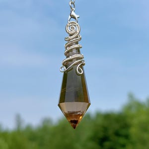 Może przedstawiać: Fasetowany, brązowy kryształowy wahadło z owinięciem z srebrnego drutu. Kryształ jest zawieszony na srebrnym łańcuszku i zapięciu. Tło to rozmyty widok niebieskiego nieba i zieleni.