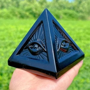 May include: Black obsidian pyramid with two carved eyes on each side. The pyramid is polished and has a glossy finish.
