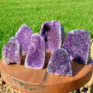 May include: A collection of amethyst crystal geodes in varying shapes and sizes. The geodes display a rich purple color with crystalline structures. They are arranged on a round, wooden surface, with a green grassy background.