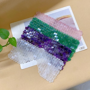 Könnte beinhalten: Vier Augenmasken aus verschiedenen Edelsteinen. Die Masken sind übereinander gestapelt, wobei die weiße Maske unten liegt, gefolgt von den violetten, grünen und rosa Masken. Die Masken bestehen aus glatten, polierten Steinen und haben eine rechteckige Form.