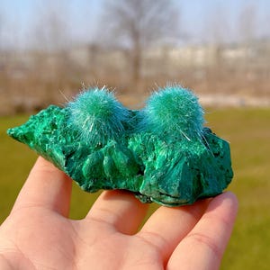 May include: A green malachite rock with two clusters of light green, fuzzy crystals on top.