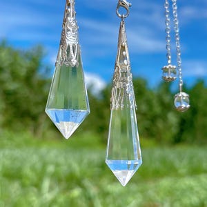 Puede incluir: Colgantes de cristal transparente y facetado con adornadas tapas y cadenas de metal plateado. Los colgantes tienen forma de lágrima, reflejando el cielo azul y el follaje verde del fondo. Los cristales están suspendidos, creando una estética delicada y elegante.