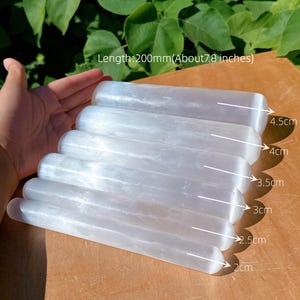 May include: Six white selenite wands of varying lengths, the longest being approximately 200mm. Each wand has a rounded end. The wands are arranged on a wooden surface, with green leaves visible in the background.