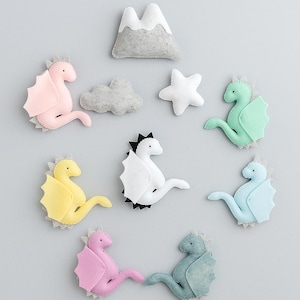 May include: A collection of felt dragon ornaments in various pastel colors, including pink, yellow, and blue, arranged with a white star, a gray cloud, and a mountain. These whimsical decorations are perfect for a nursery or children's room.